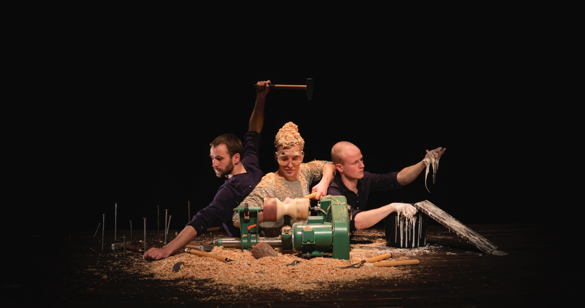 Drei Männer arbeiten an einer Drehbank und sind mit Holzspänen bedeckt, während einer ein Holzstück hält und ein anderer nasse Flüssigkeit aus einem Eimer hebt.