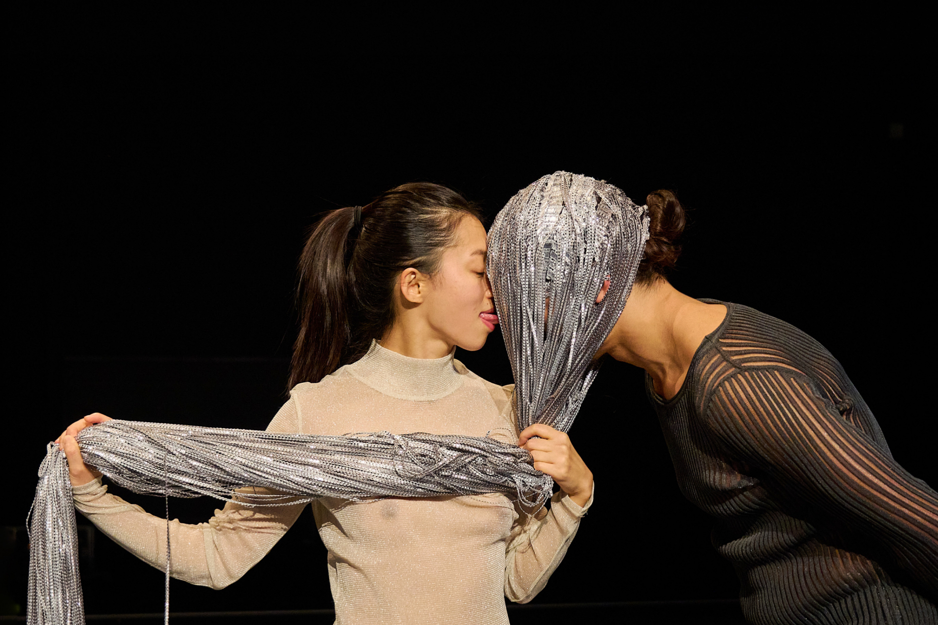 Zwei Personen stehen sich gegenüber, wobei eine Person einen silbernen Schleier hält und ihr Gesicht verdeckt, während die andere den Schleier mit der Zunge berührt.