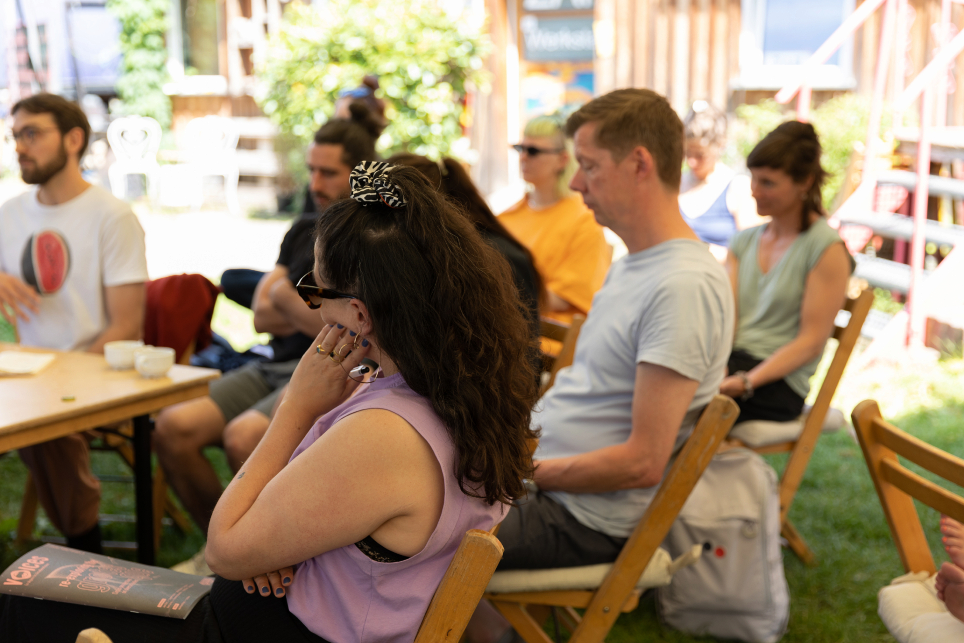 Eine Gruppe von Menschen sitzt draußen auf Holzstühlen und hört aufmerksam zu.